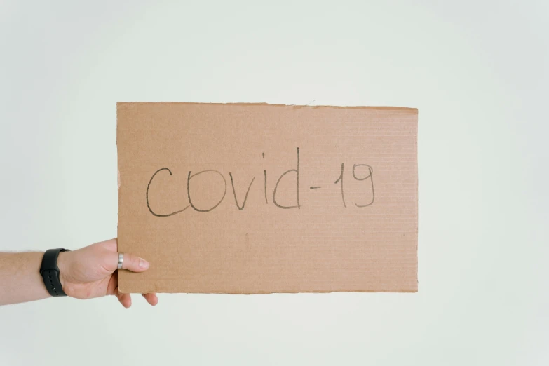 a person holding a cardboard sign with the word covidi - 19 written on it, an album cover, trending on pexels, healthcare, background image, minimalissimo, ooak