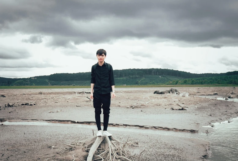 a man standing on top of a tree stump, an album cover, inspired by Xia Yong, unsplash, androgynous person, shoreline, overcast mood, heath clifford