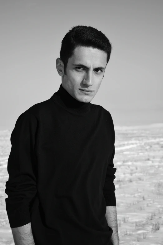 a black and white photo of a man standing in the snow, a black and white photo, inspired by Alexis Grimou, les nabis, black turtle neck shirt, in the desert beside the gulf, a handsome man，black short hair, kafka