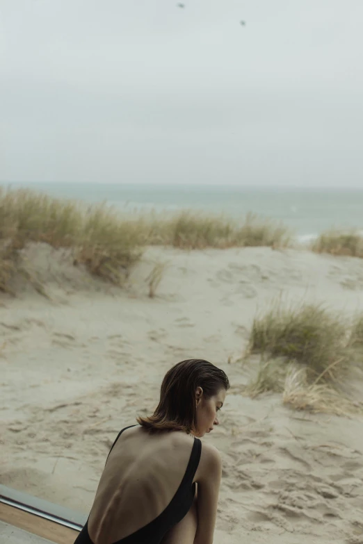 a woman sitting on a bench looking out a window at the beach, by Caro Niederer, unsplash, conceptual art, girl walking between dunes, photo of emma watson in swimsuit, denmark, medium format. soft light