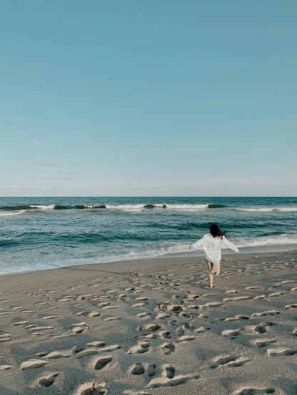 a woman standing on top of a sandy beach next to the ocean, walking away from the camera, wearing a simple robe, new jersey, unsplash 4k