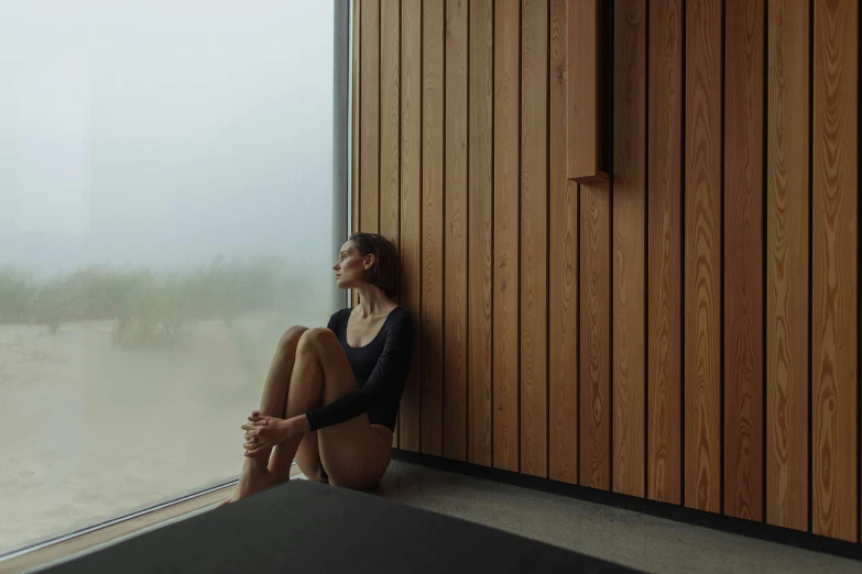 a woman sitting on a window sill looking out a window, by Emma Andijewska, minimalism, sauna, sand mists, floor to ceiling window, in a beachfront environment