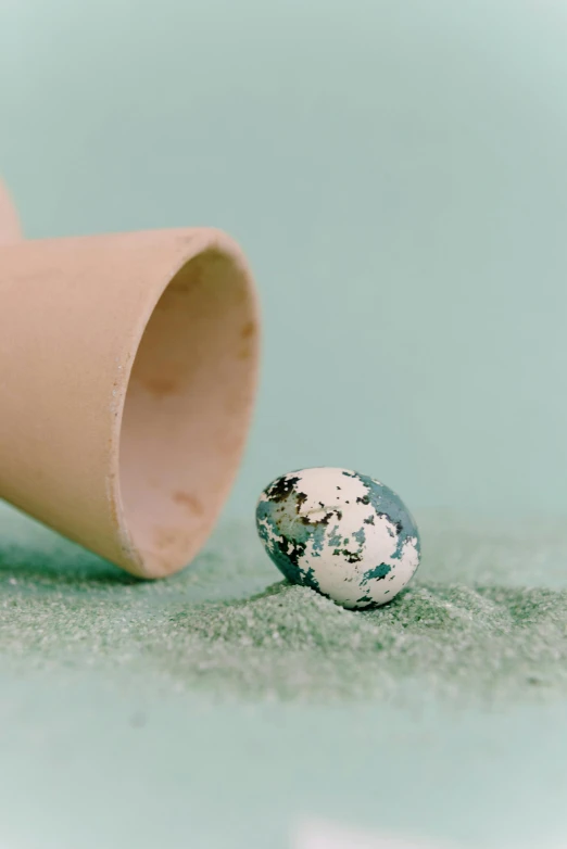 an egg sitting on top of a table next to a cup, an album cover, by Elsa Bleda, unsplash, magic realism, sand, green pupills, made of liquid metal and marble, white cyan