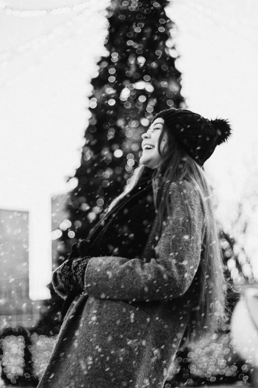 a woman standing in front of a christmas tree, a black and white photo, by Ai-Mitsu, pexels contest winner, realism, snow fall, laughing, snowstorm ::5, (beautiful) girl