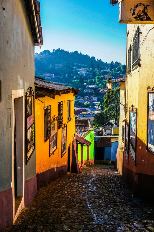 a narrow cobblestone street with a mountain in the background, an album cover, pexels contest winner, quito school, colorful house, jamaica, square, late afternoon light