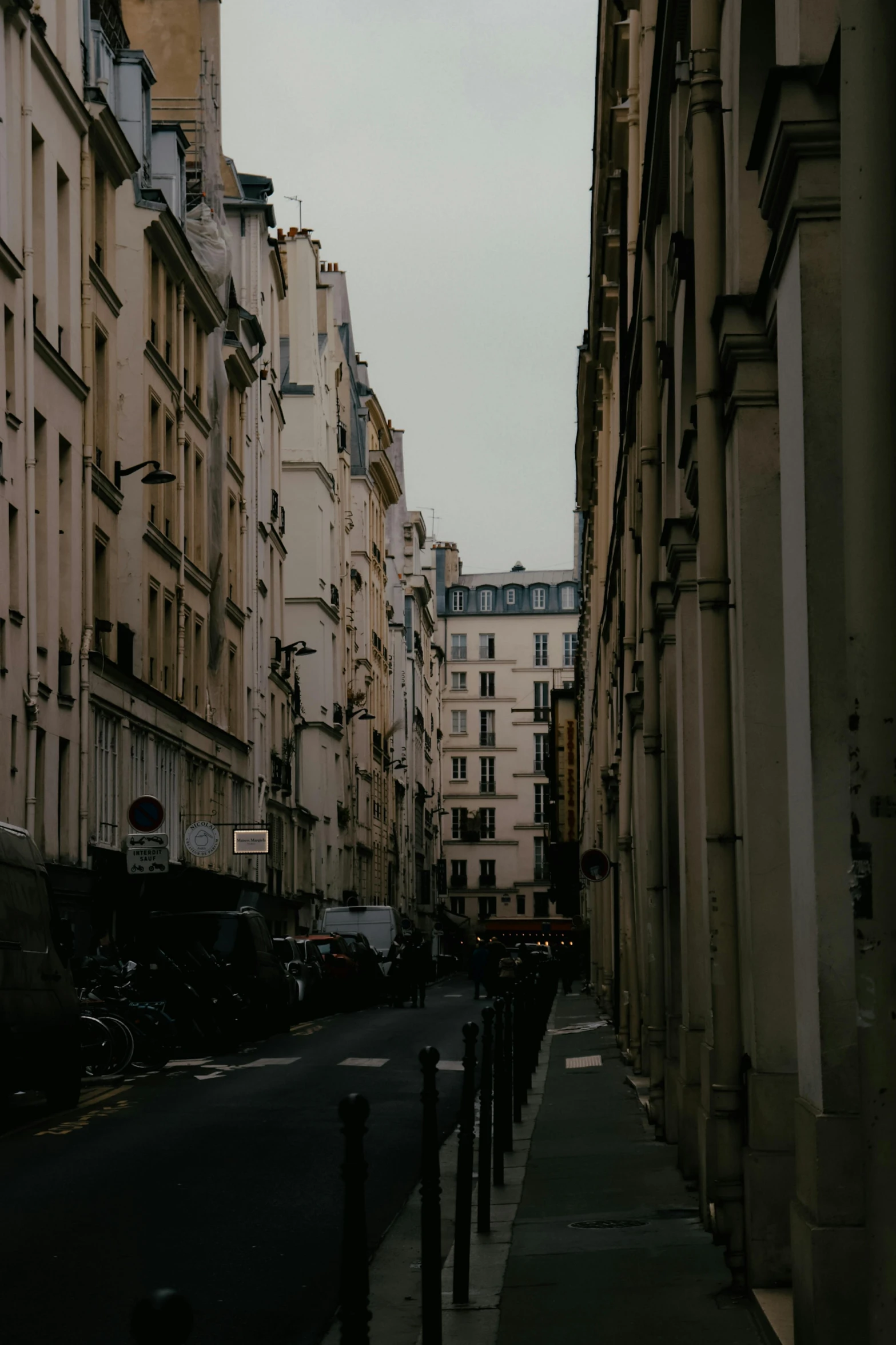 a narrow city street lined with tall buildings, a photo, pexels contest winner, paris school, barely visible from the shadows, nice slight overcast weather, neighborhood, gothic city streets behind her