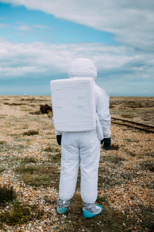 a person in a white suit standing in a field, inspired by Derek Jarman, unsplash, wearing a space suit, on the coast, on a landing pad, back facing