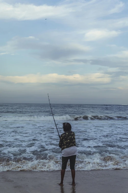 a woman standing on top of a beach next to the ocean, fishing, rough waters, in the evening, afternoon