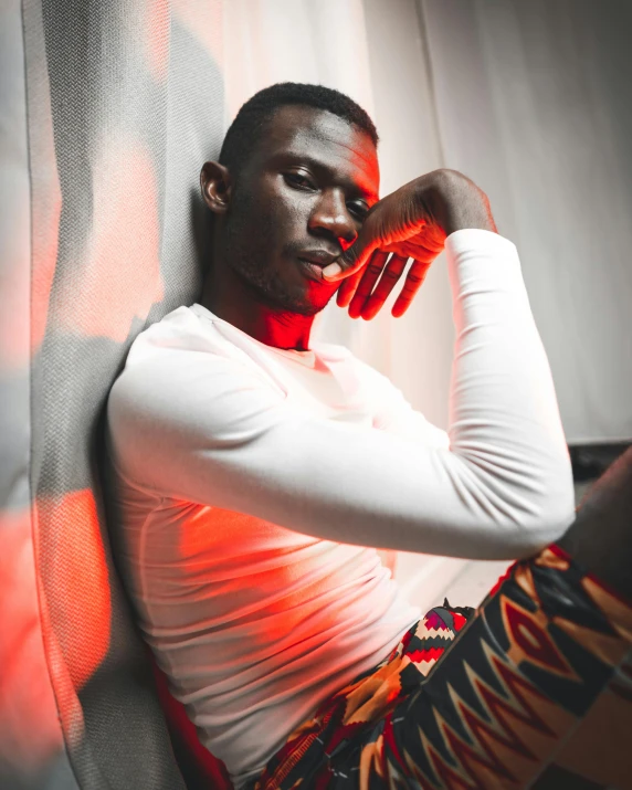 a man in a white shirt leaning against a wall, inspired by David Bailly, trending on pexels, afrofuturism, bisexual lighting, sitting on a bed, in a glowing skirt, red cloth around his shoulders