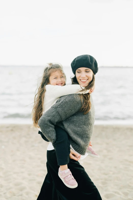 a woman carrying a little girl on the beach, by Anita Malfatti, unsplash, wearing a sweater, on white background, smiling slightly, with black beanie on head