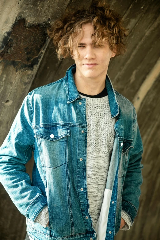 a young man standing in front of a stone wall, a picture, by Jacob Toorenvliet, trending on pexels, light brown messy hair, denim jacket, hollywood promotional image, male teenager