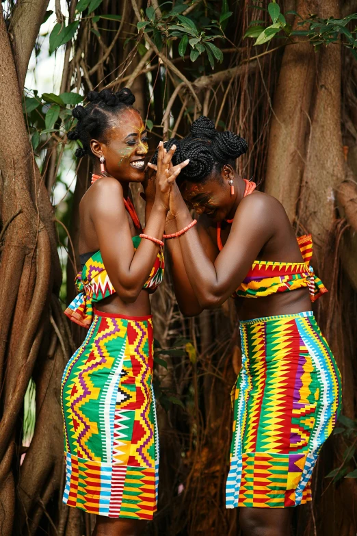 a couple of women standing next to a tree, an album cover, inspired by Ras Akyem, pexels contest winner, happening, colorful uniforms, loin cloth, twins, celebration