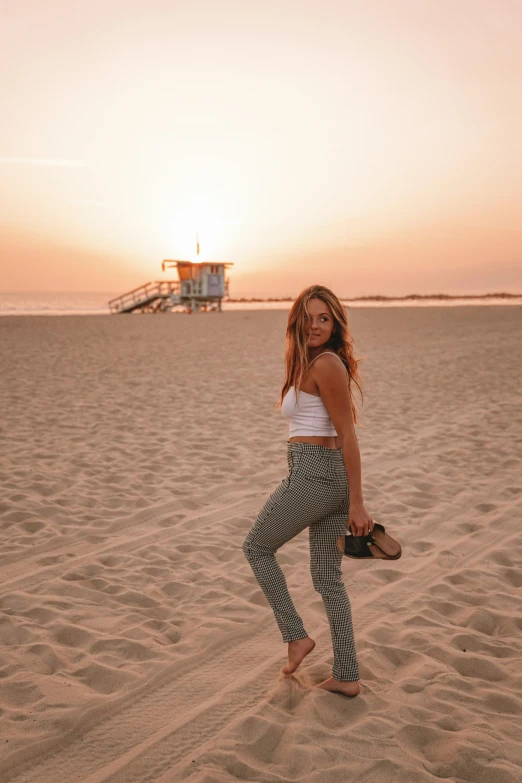 a woman standing on top of a sandy beach, by Robbie Trevino, wearing cargo pants, hollywood standard, at beach at sunset, chloe bennet