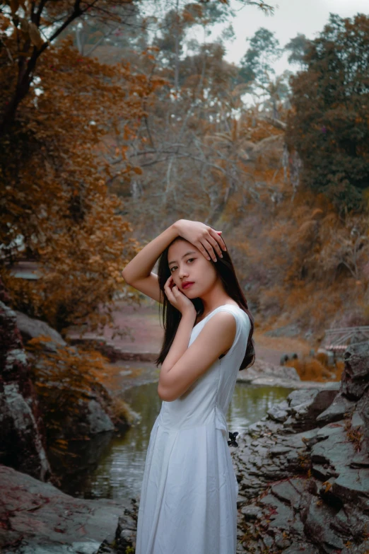 a woman in a white dress standing next to a river, inspired by Elsa Bleda, pexels contest winner, realism, young asian girl, muted fall colors, ((portrait)), medium format