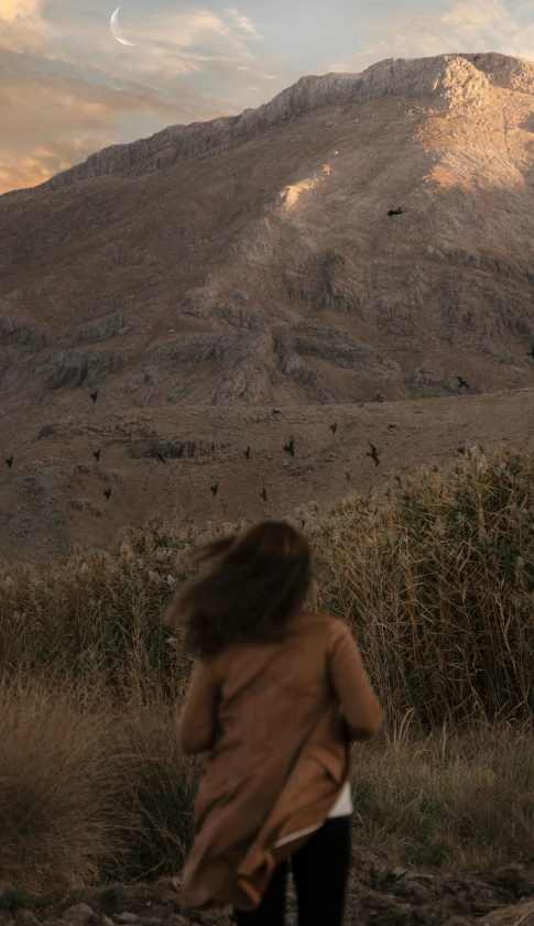 a woman standing in a field with a mountain in the background, pexels contest winner, naturalism, tribe members chasing, beige and dark atmosphere, brown, in a volcano