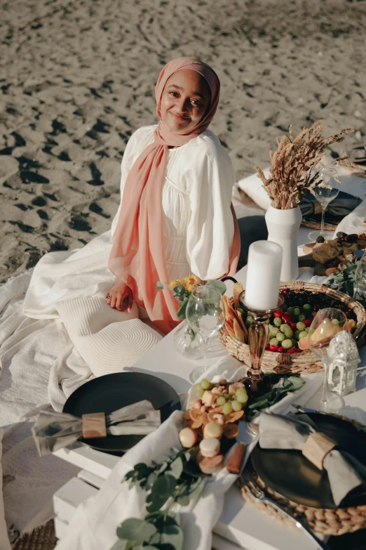 a woman sitting at a picnic table on the beach, inspired by Modest Urgell, pexels contest winner, renaissance, white hijab, overflowing feast buffet table, smiling down from above, dressed thobe
