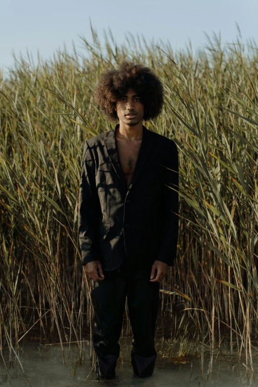 a man standing in a body of water next to tall grass, an album cover, renaissance, afro hair, a man wearing a black jacket, looking towards camera, posing on wheat field