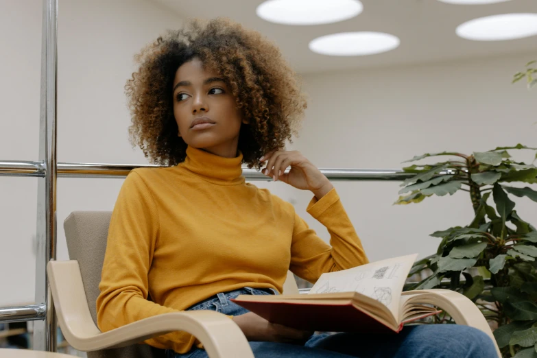 a woman sitting in a chair reading a book, a portrait, trending on pexels, long afro hair, yellow clothes, realistic », at college