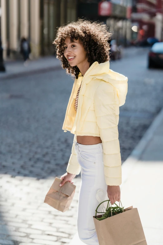 a woman walking down the street carrying a shopping bag, by Nina Hamnett, trending on pexels, happening, wavy hair yellow theme, model wears a puffer jacket, bare midriff, white puffy outfit