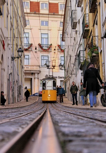a yellow train traveling down a street next to tall buildings, pexels contest winner, renaissance, lisbon, gray men, youtube thumbnail, people watching around