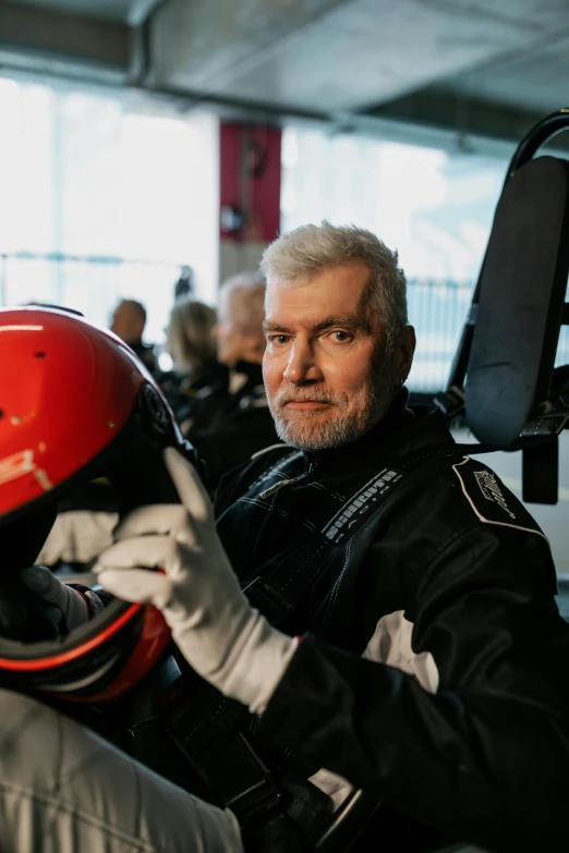 a man sitting in the driver's seat of a race car, wearing flight suit, dark grey haired man, formula 1 garage, zero gravity