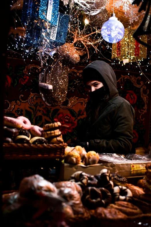 a man standing in front of a table filled with doughnuts, by Julia Pishtar, wearing a dark hood, festive, balaclava, market setting