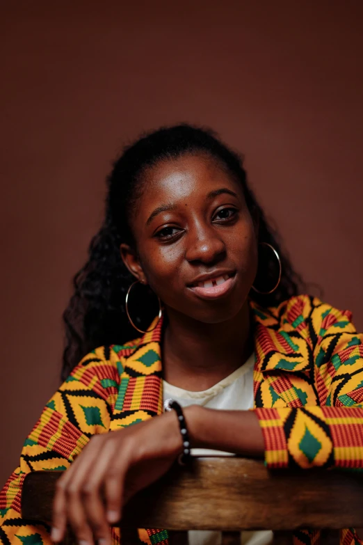 a woman sitting on top of a wooden chair, by Chinwe Chukwuogo-Roy, trending on unsplash, hurufiyya, wearing jacket, headshot portrait, patterned, ( ( brown skin ) )