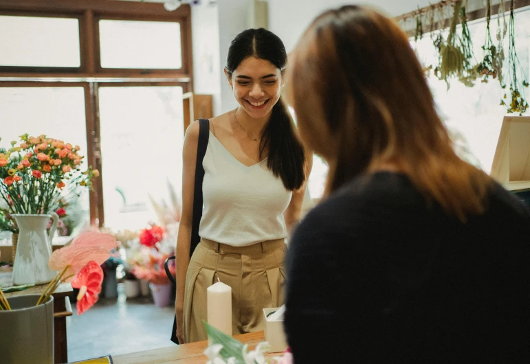 a woman talking to another woman in a flower shop, pexels contest winner, slightly awkward smile, a person standing in front of a, promo image, on a white table