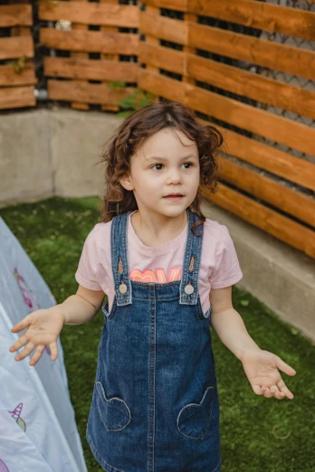 a little girl that is standing in the grass, wearing blue jean overalls, shrugging arms, softplay, epk