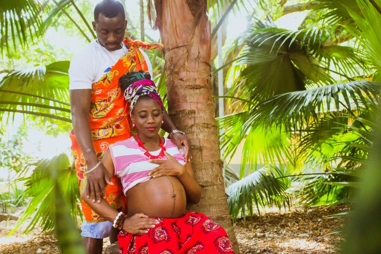 a man and a woman standing next to a palm tree, by Ingrida Kadaka, pexels, symbolism, pregnant belly, yoruba body paint, laying under a tree on a farm, avatar image