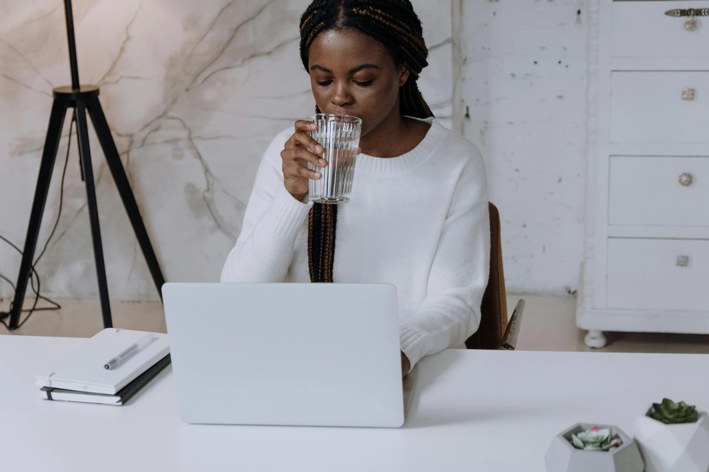 a woman sitting at a table with a laptop and a glass of water, trending on pexels, wearing a white sweater, avatar image, black female, pokimane