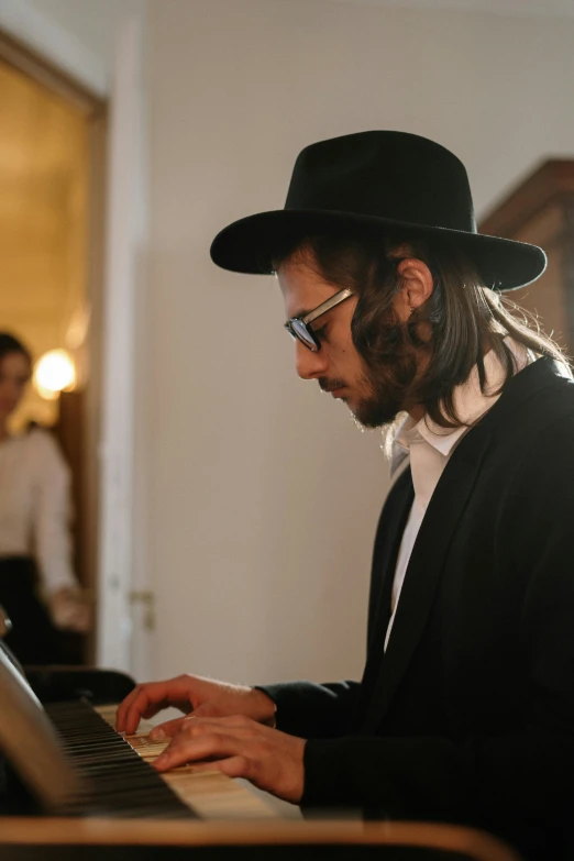 a man in a hat playing a piano, by Elias Goldberg, trending on unsplash, jewish young man with glasses, elegantly dressed, official screenshot, trending on artist