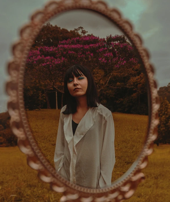 a woman standing in front of a mirror in a field, an album cover, pexels contest winner, flowers in her dark hair, looking to camera, high quality photo, gold framed