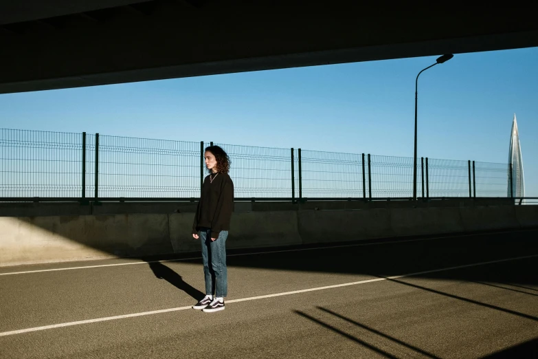 a woman standing in a parking lot next to a fence, by Nathalie Rattner, finn wolfhard, high quality photo, overpass, slightly minimal