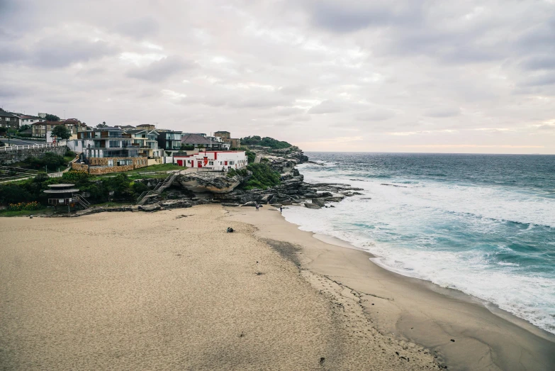 a sandy beach next to the ocean under a cloudy sky, by Elsa Bleda, pexels contest winner, bondi beach in the background, the village on the cliff, hazy and dreary, conde nast traveler photo