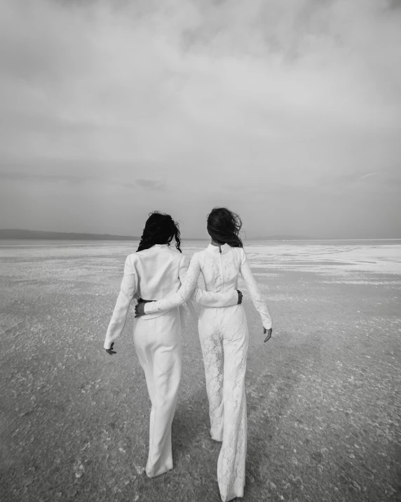 two women walking on a beach holding hands, a black and white photo, by Lucia Peka, unsplash, wearing futuristic white suit, lgbt, white salt, twins