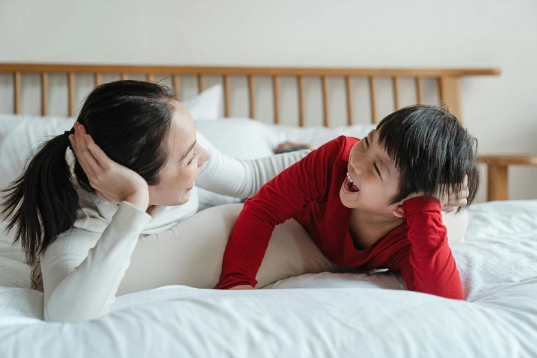 a woman laying on top of a bed next to a child, pexels contest winner, smiling at each other, asian male, shouting, wearing red clothes