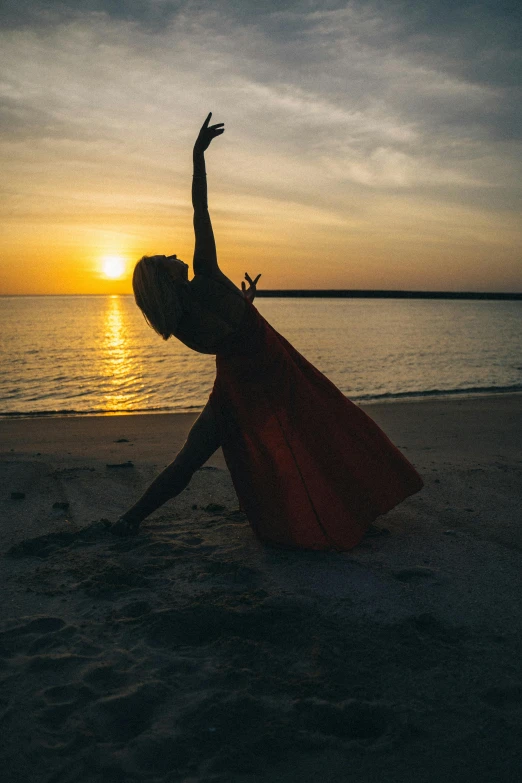 a woman in a red cape on the beach, pexels contest winner, arabesque, yoga pose, sun setting, ballet pose, grainy