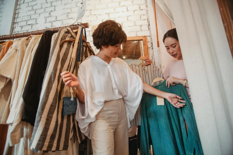 a woman standing in front of a rack of clothes, trending on pexels, renaissance, woman holding another woman, inspect in inventory image, white and teal garment, cottagecore hippie