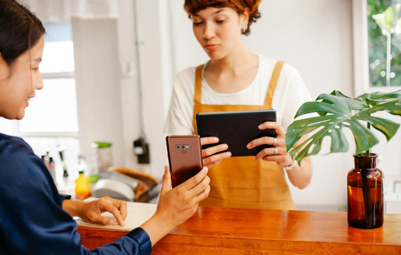 two women sitting at a table looking at a tablet, trending on pexels, realism, cash register, holding a very advance phone, a wooden, avatar image