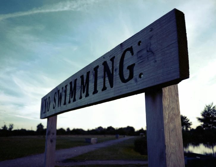 a wooden sign sitting on the side of a road, by Bob Ringwood, unsplash, summer swimming party, 2000s photo, early evening, partly sunken! in the lake!