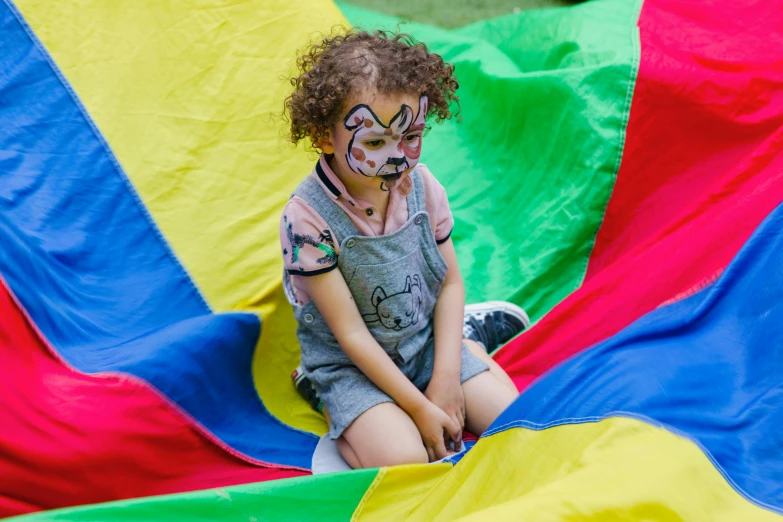 a little girl with face paint sitting on a parachute, by Arabella Rankin, pexels contest winner, process art, square, activity play centre, pride flag in background, toddler