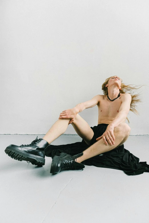 a shirtless man sitting on the floor in front of a white wall, an album cover, by Grace Polit, trending on pexels, visual art, portrait of kim petras, heavy boots, captures emotion and movement, attractive androgynous humanoid