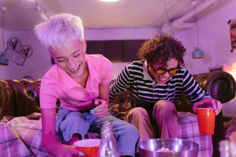 a couple of women sitting on top of a couch, by Nick Fudge, trending on pexels, happening, purple and pink and blue neons, bowl filled with food, teenage boy, laughing