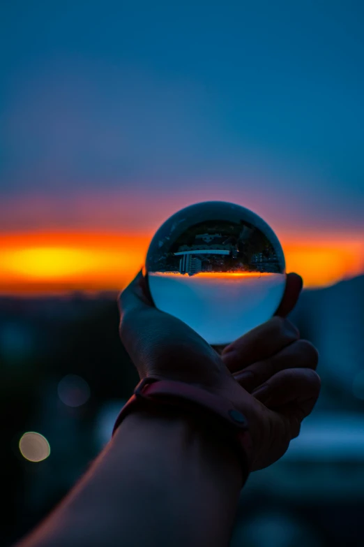 a person holding a crystal ball with a sunset in the background, a picture, by Doug Ohlson, pexels contest winner, city views, glass cover, panoramic photography, water droplets frozen in time