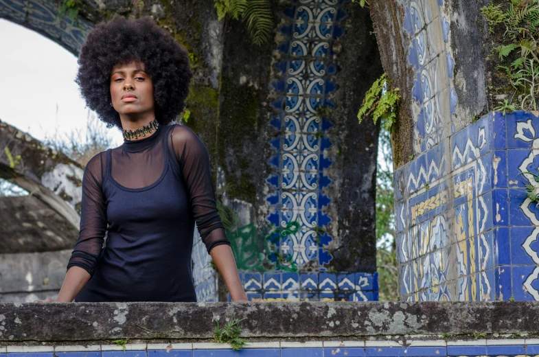 a woman standing on top of a blue and white tiled wall, an album cover, inspired by Almeida Júnior, pexels contest winner, afrofuturism, black curly hair, lush surroundings, las pozas, ancient catedral behind her