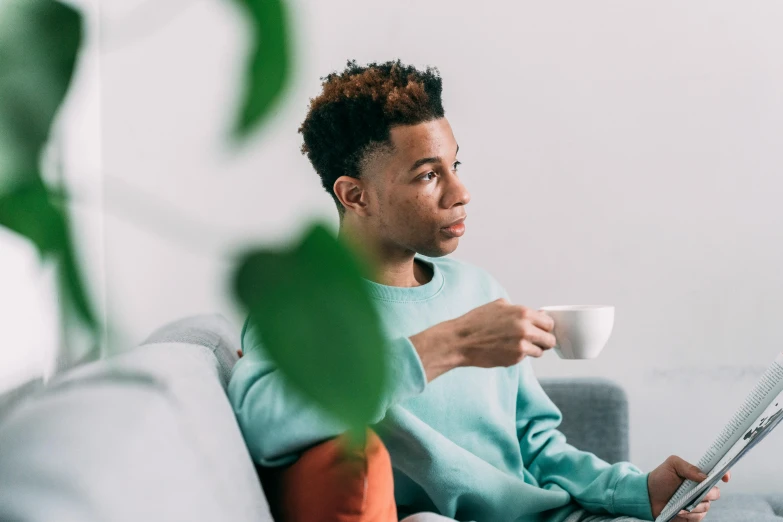 a man sitting on a couch reading a newspaper and drinking a cup of coffee, by Sebastian Vrancx, trending on pexels, black teenage boy, wearing a green sweater, avatar image, drinking tea