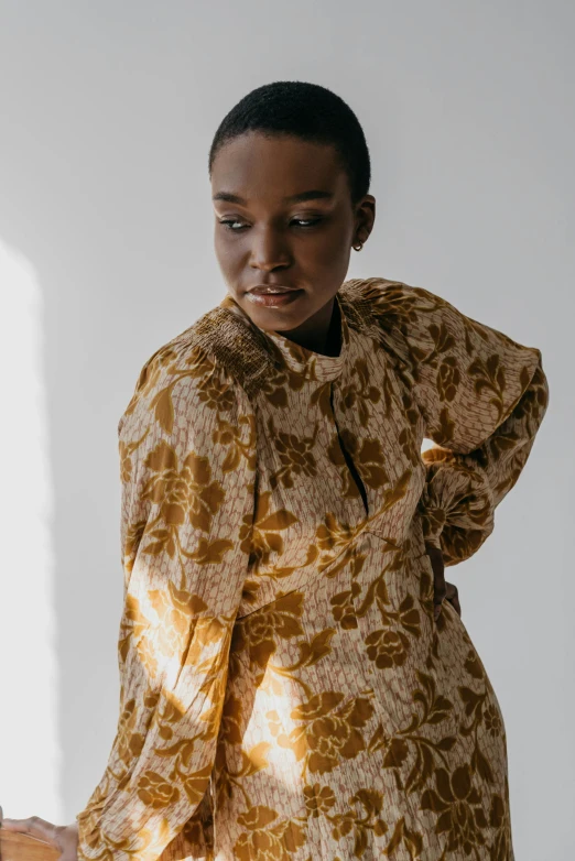 a woman standing in front of a window, by Winona Nelson, wearing yellow floral blouse, beige and gold tones, maria borges, muted brown