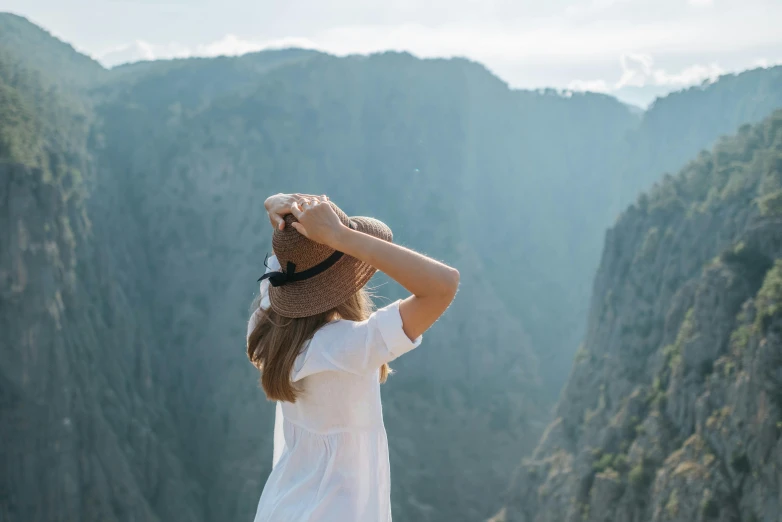 a woman standing on top of a mountain with a hat on, pexels contest winner, wearing a linen shirt, view from side, sharp cliffs, her hair is tied above her head