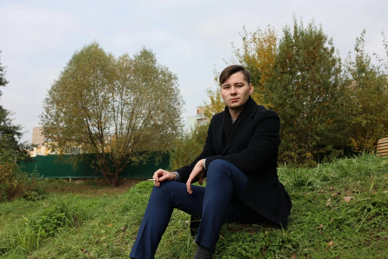 a man sitting on top of a lush green hillside, an album cover, realism, julian ope, he is wearing a black trenchcoat, in a city park, photographed for reuters